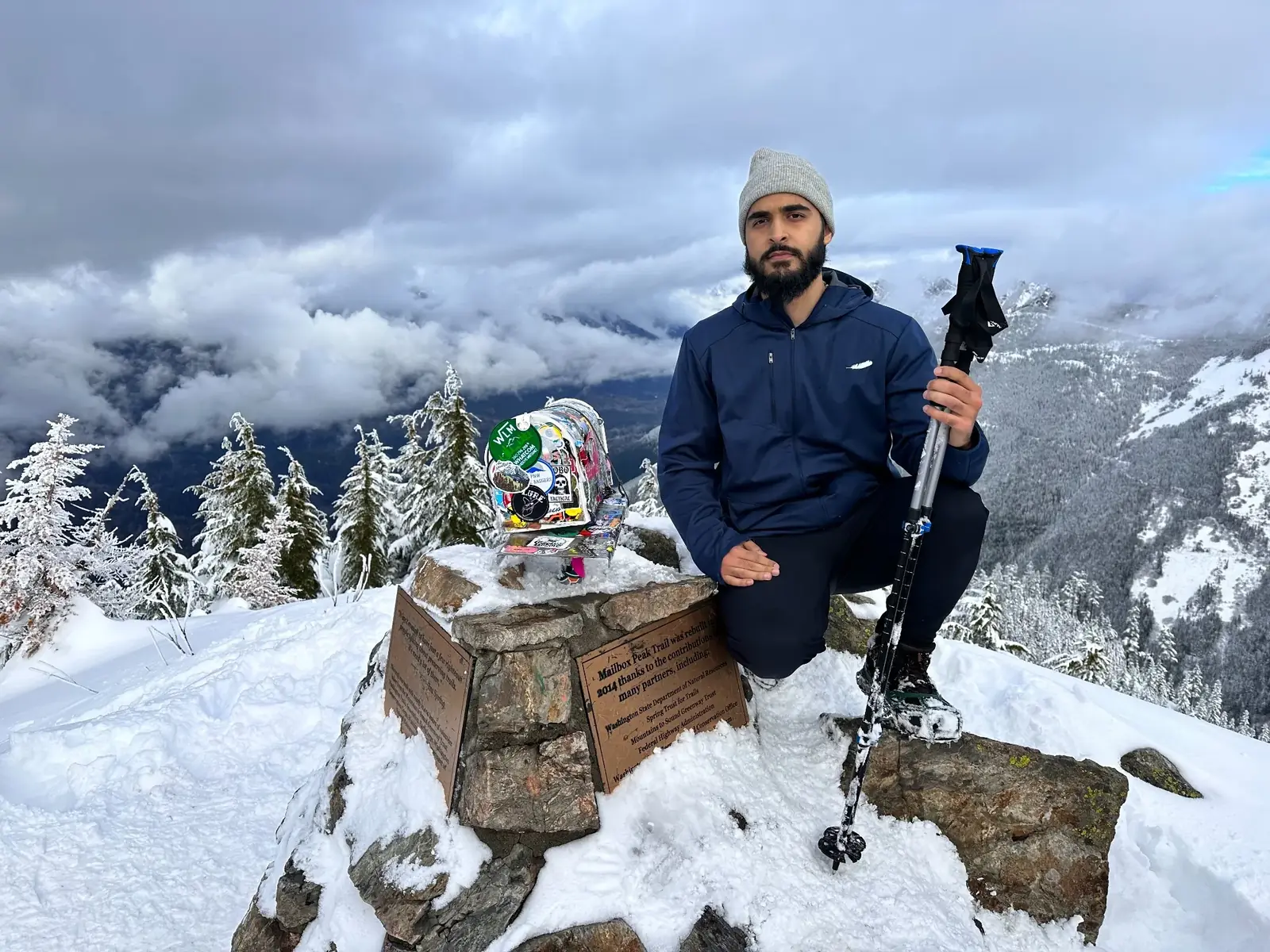 Waleed kneeling beside the summit mailbox on a snowy mountain