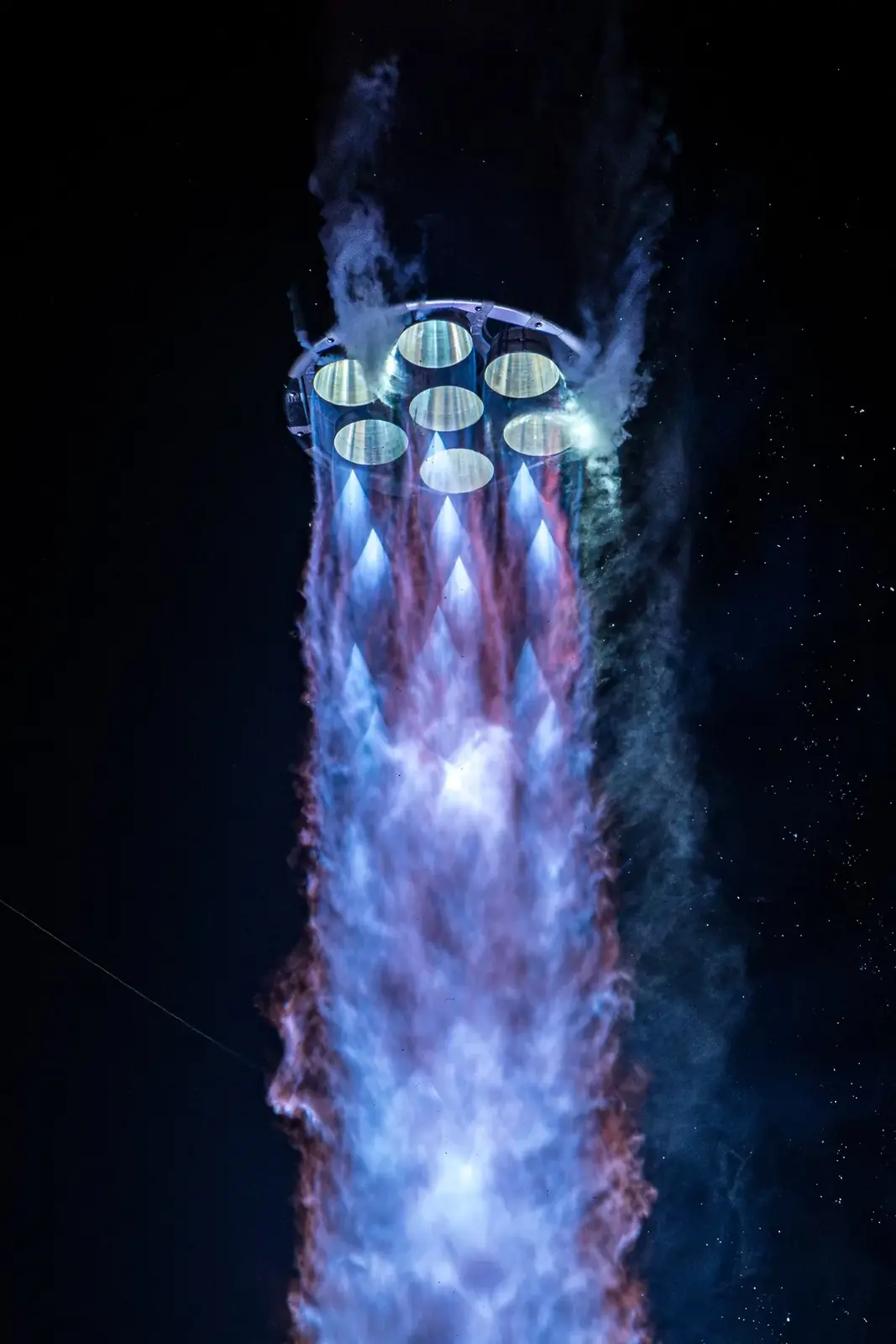 Close view beneath a rocket showing seven engine nozzles and a blue-purple exhaust plume